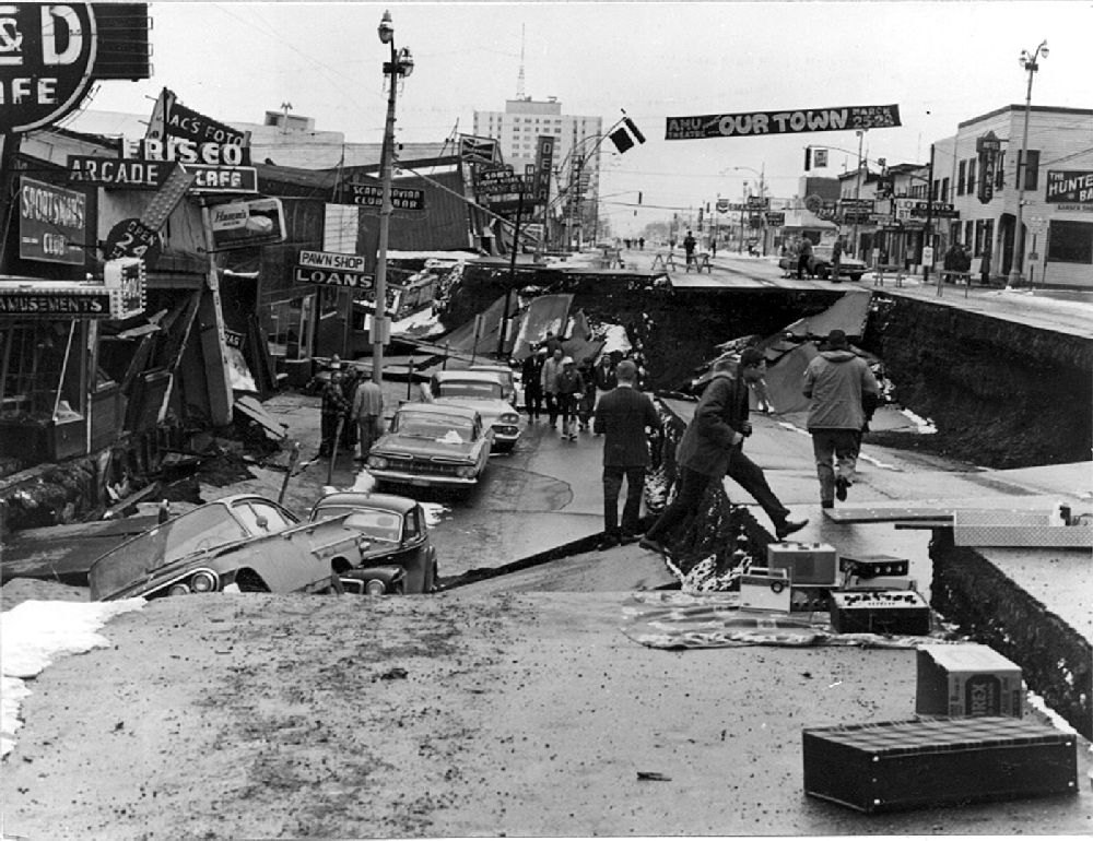 The street is sunk in with people walking on different levels. Cars and buildings are trapped and slanted. Damage/broken material scattered.