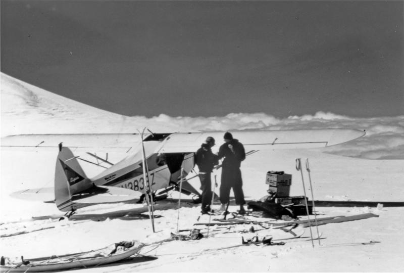 Two persons next to a plane and equipment on tundra snow