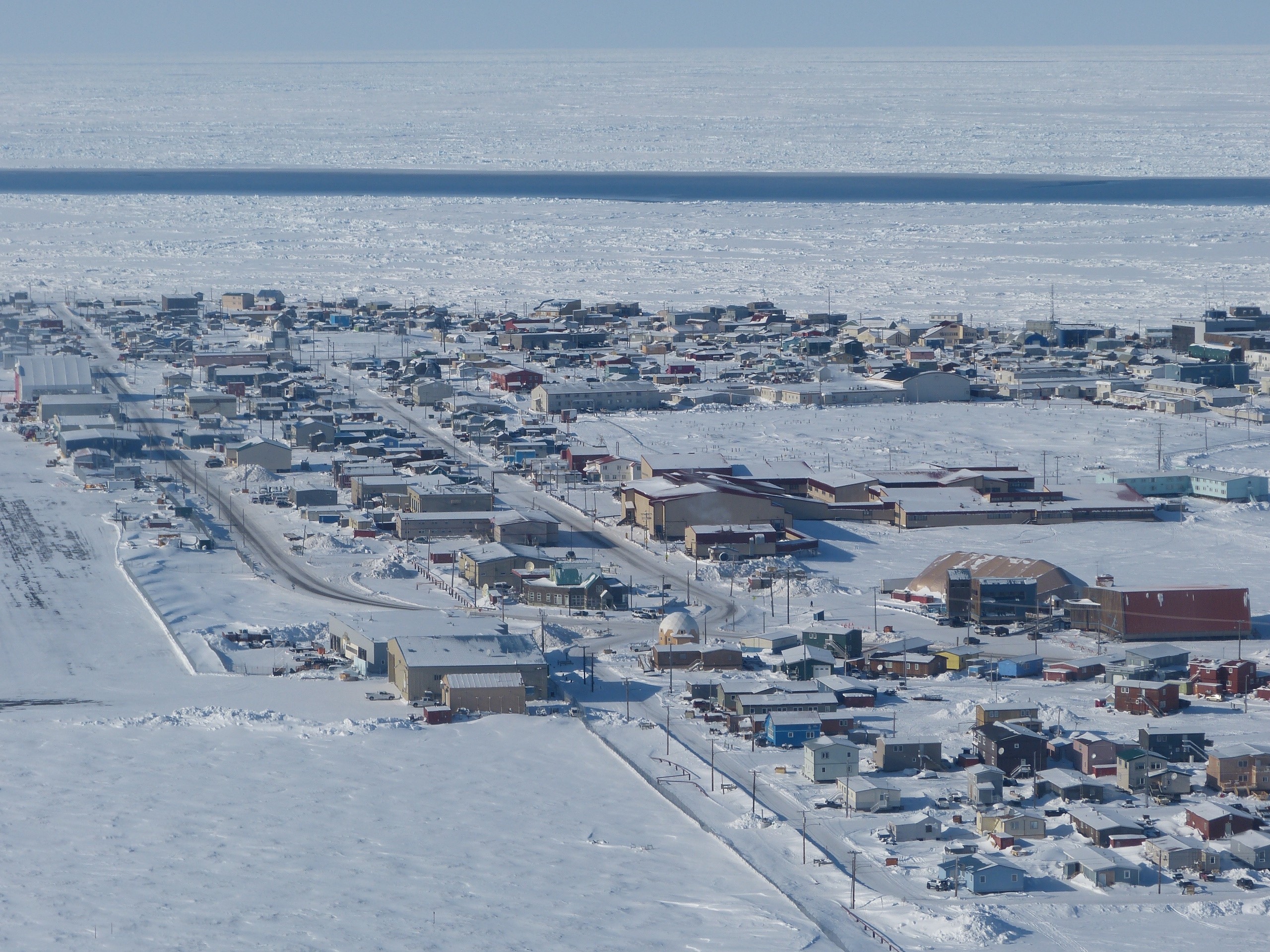 Aerial view of Utqiaġvik, Alaska, showing a snow-covered coastal town bordered by sea ice. A long, narrow strip of open water called an open flaw lead cuts across the frozen ocean, separating the solid landfast ice attached to shore from the drifting pack ice offshore. Photo by Andrew Mahoney