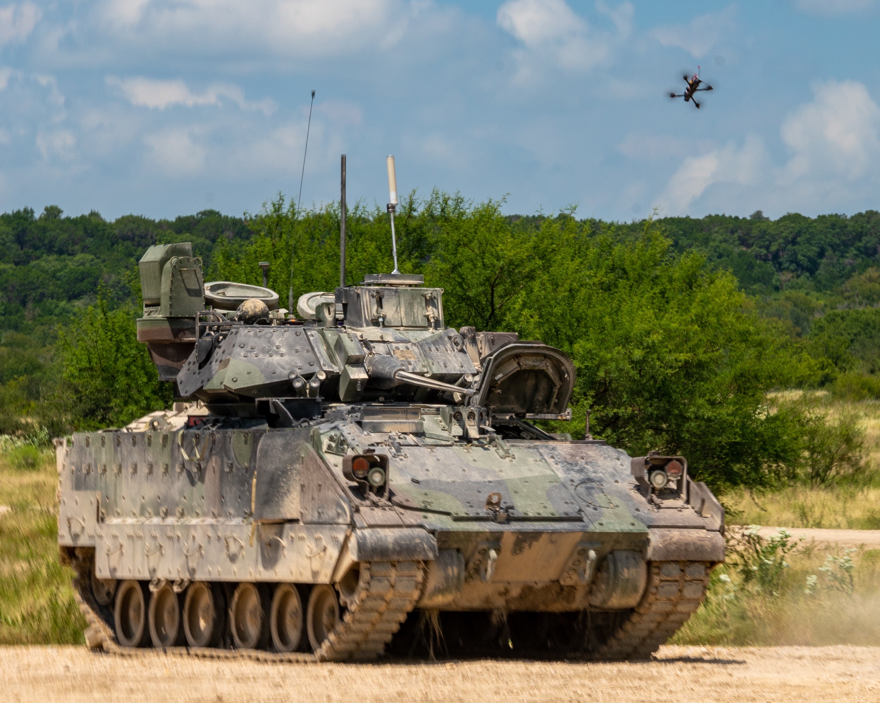 A small quadcopter unmanned aircraft system hovers above an M2 Bradley Infantry Fighting Vehicle on a dirt training range at Fort Hood, Texas, with trees in the background. The scene, captured Aug. 27, 2025, during Operation Return of the Condor, shows the drone positioned overhead in a test.