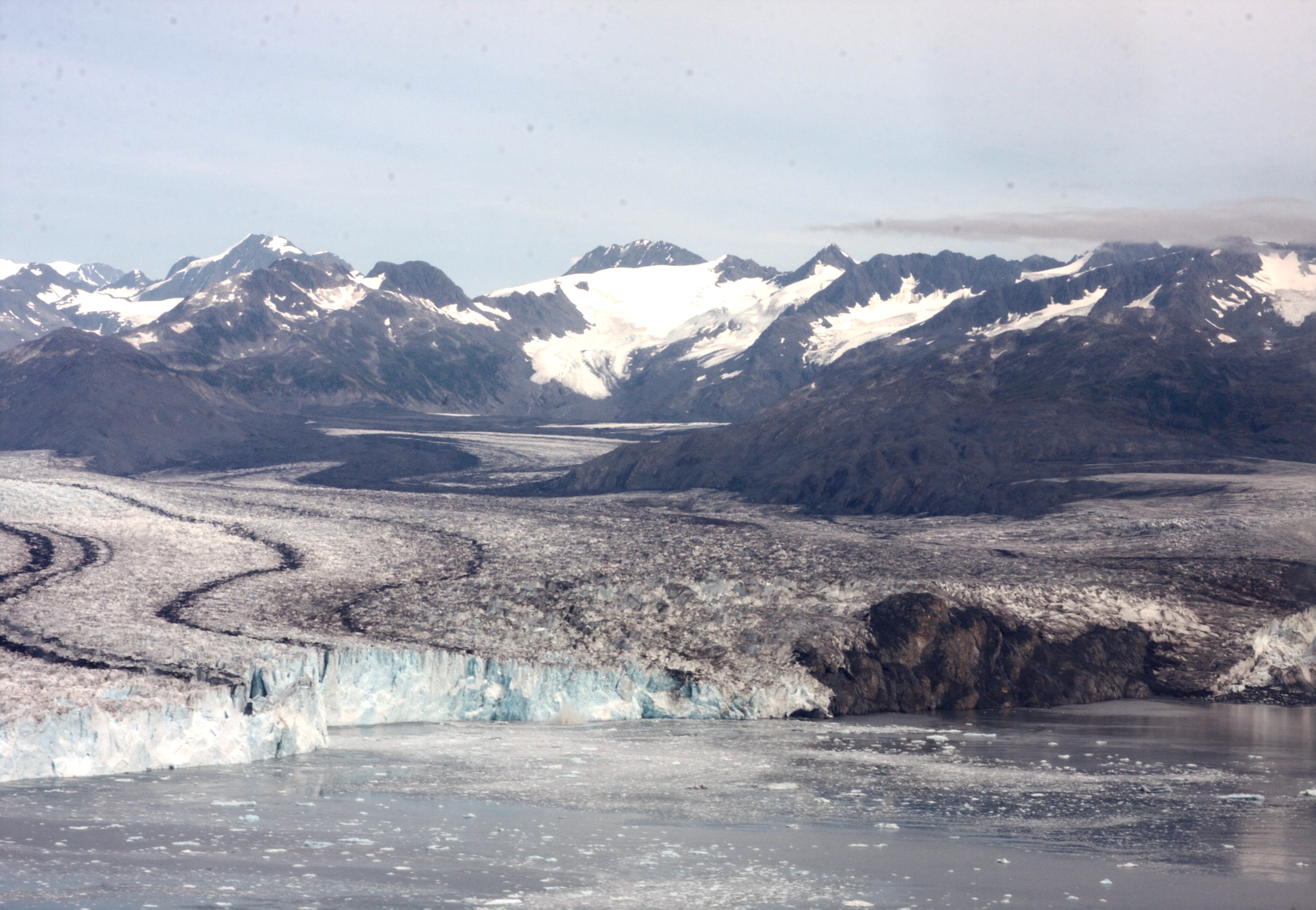 A wide view of Columbia Glacier in Alaska shows a broad river of ice flowing between dark, rocky mountains, with snow-covered peaks in the distance and floating ice in the water at the glacier’s front. 