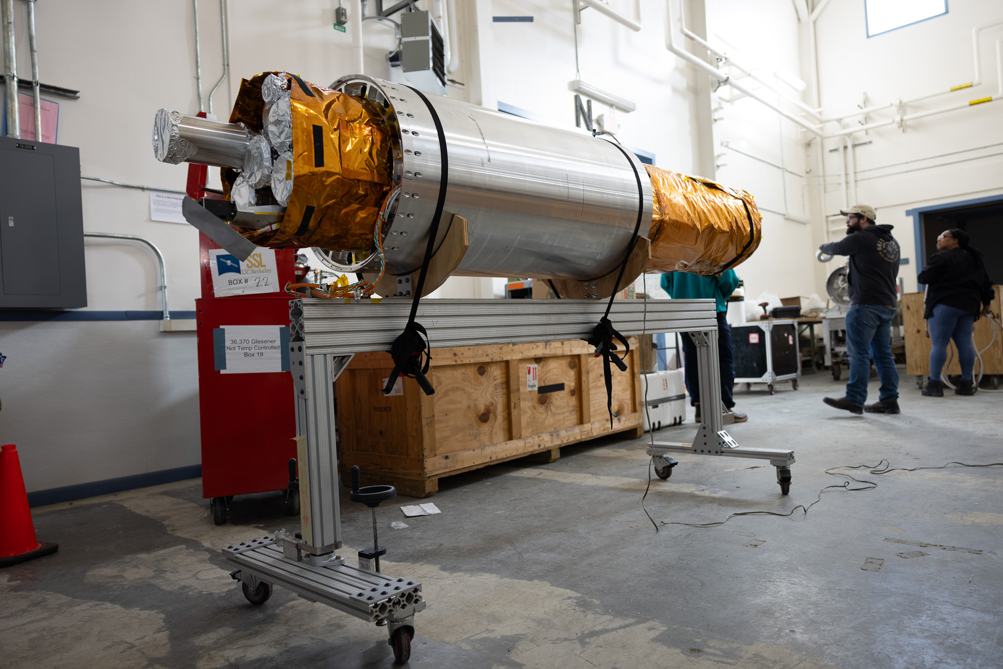 The payload section of the FOXSI-5 mission rests on a wheeled support frame inside the payload assembly building at Poker Flat Research Range. The cylindrical instrument, wrapped partly in gold thermal insulation, is surrounded by equipment crates and technicians preparing it for flight. 