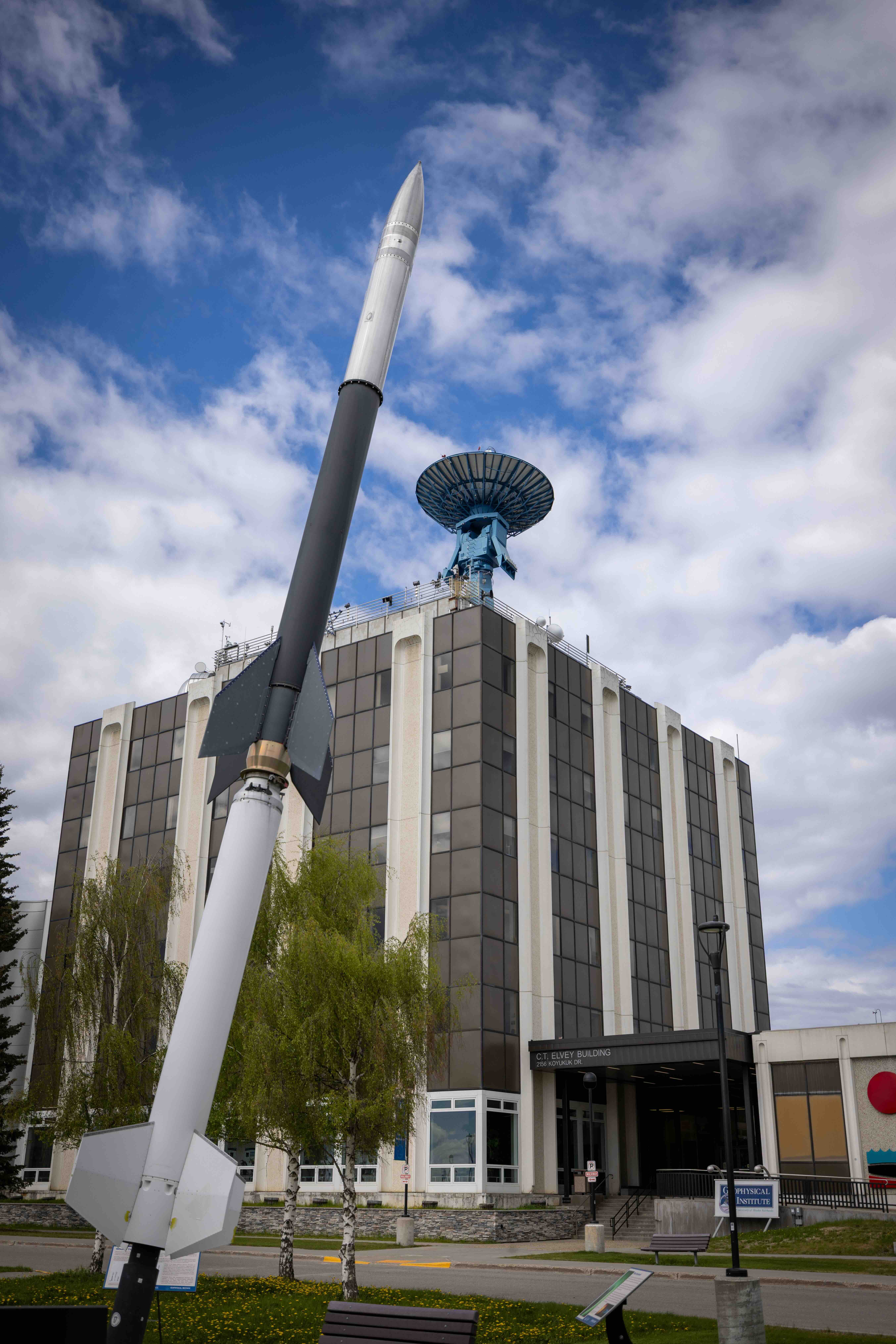 Visitors on the Geophysical Institute summer tour will learn about the large blue satellite antenna atop the Elvey Building and the sounding rocket installation displayed on the lawn outside. The photo shows the rocket model rising in the foreground in front of the building under a partly cloudy sky