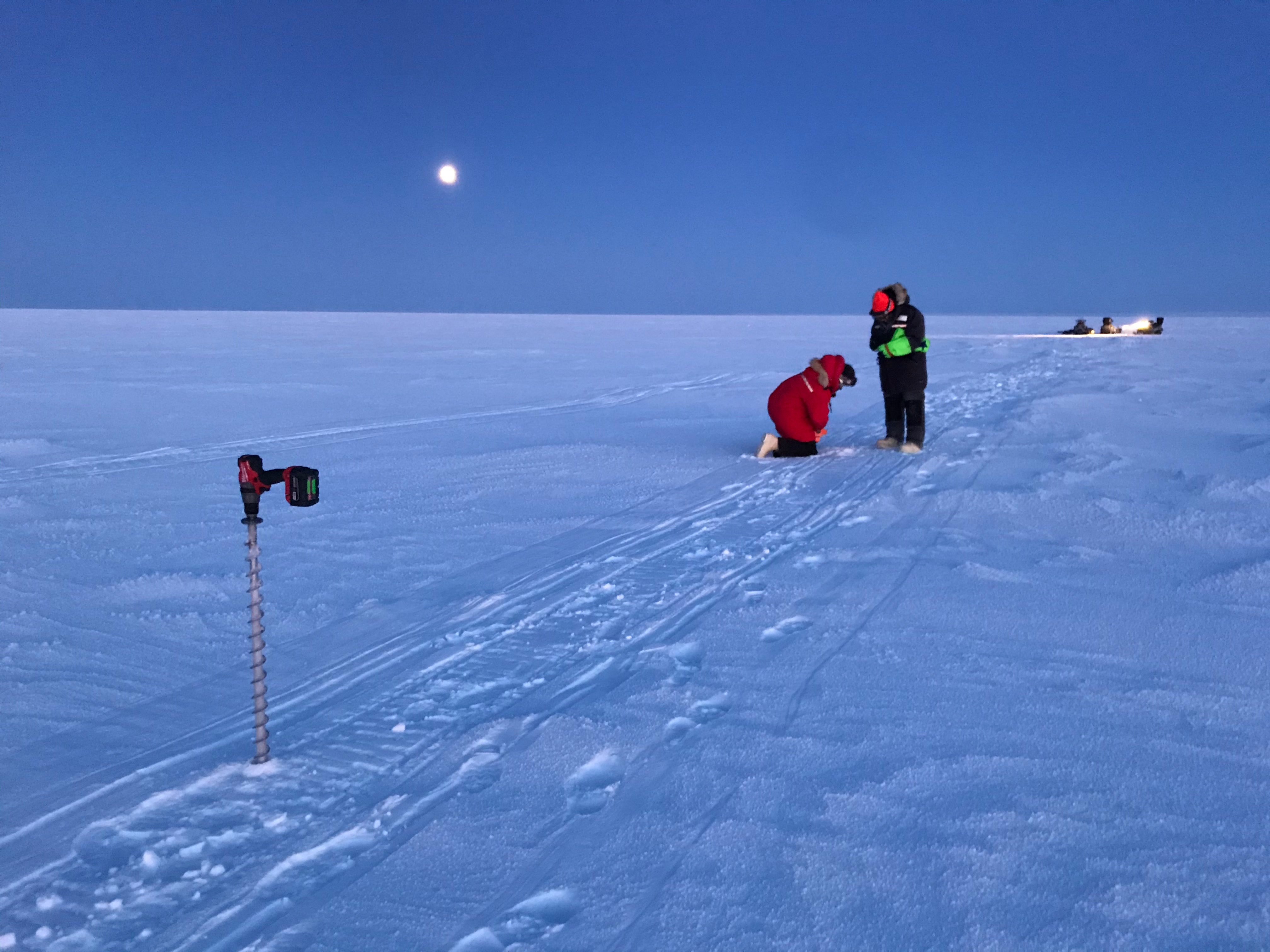  Two researchers in heavy winter clothing kneel and stand on the snow-covered surface of Teshekpuk Lake as they conduct ice measurements under a moonlit Arctic sky. An ice auger stands in the foreground near snowmachine tracks, while additional team members and equipment are in the distance. 