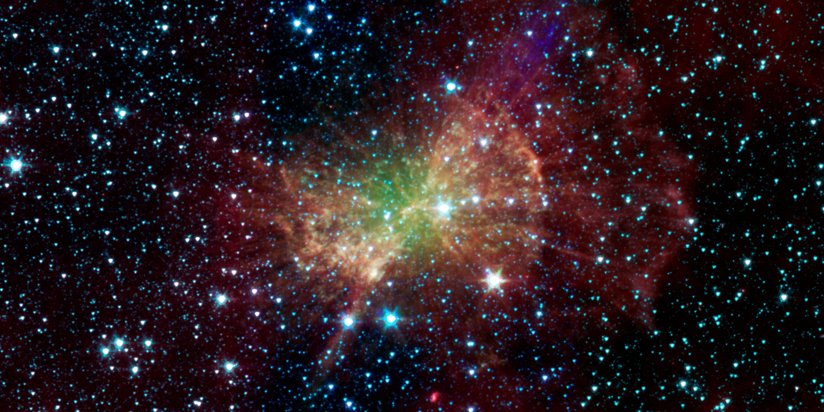 The Dumbbell Nebula glows in infrared light in this 2011 image from NASA’s Spitzer Space Telescope. A broad, irregular cloud of green, yellow and orange gas fills the center, surrounded by wispy red clouds against the blackness of space. Hundreds of bright blue-white stars are scattered throughout.