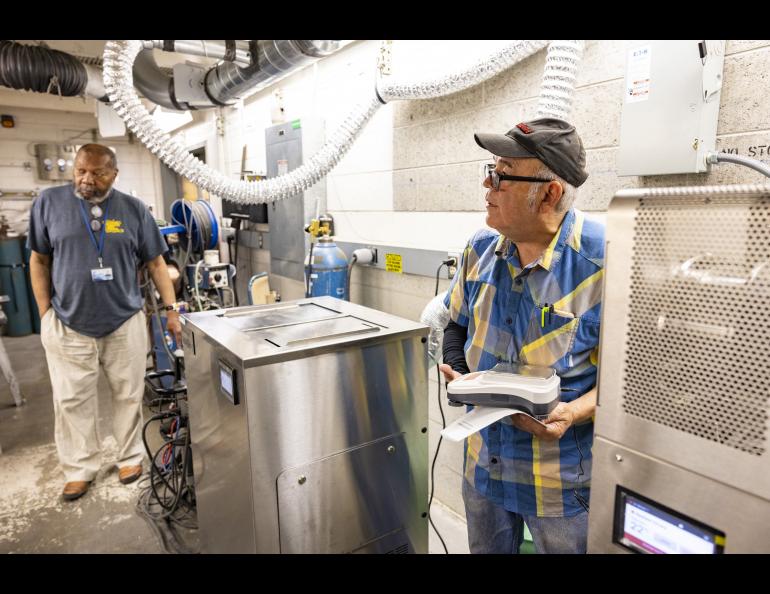 Jesse Atencio holds a scale where 3D printed objects are weighed. Objects printed on the new metal 3D printer shrink to their intended size during the washing, curing and firing process. Machine Shop Manager Greg Shipman is also pictured. Photo by JR Ancheta