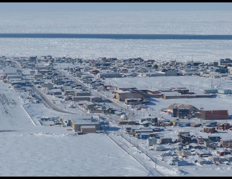 Aerial view of Utqiaġvik, Alaska, showing a snow-covered coastal town bordered by sea ice. A long, narrow strip of open water called an open flaw lead cuts across the frozen ocean, separating the solid landfast ice attached to shore from the drifting pack ice offshore. Photo by Andrew Mahoney