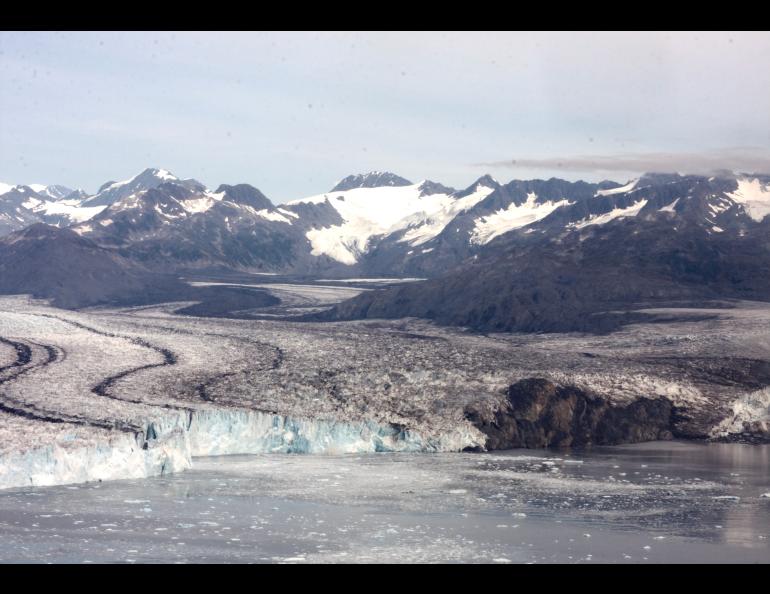 A wide view of Columbia Glacier in Alaska shows a broad river of ice flowing between dark, rocky mountains, with snow-covered peaks in the distance and floating ice in the water at the glacier’s front. 