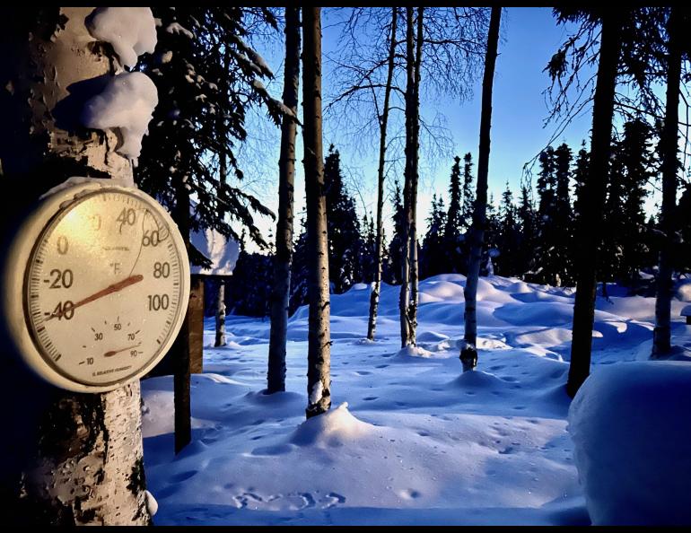 A round outdoor thermometer mounted on a snow-covered tree reads about minus 45 degrees Fahrenheit in a quiet, snow-blanketed forest of tall trees. Long blue shadows stretch across the deep snow under a clear sky. The image shows extreme cold conditions at a home in Two Rivers, Alaska.