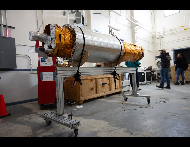 The payload section of the FOXSI-5 mission rests on a wheeled support frame inside the payload assembly building at Poker Flat Research Range. The cylindrical instrument, wrapped partly in gold thermal insulation, is surrounded by equipment crates and technicians preparing it for flight. 