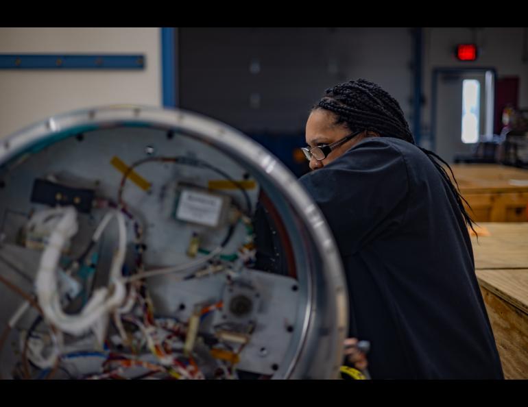 Lawanda Long, with the FOXSI-5 team, leans in to assist with payload preparation inside the payload assembly building at Poker Flat Research Range. She focuses on a cylindrical instrument in the foreground, where wiring and components are visible inside the open end.