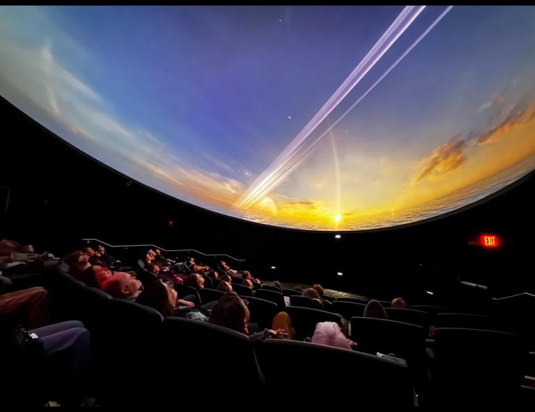 Audience members sit in reclined theater seats, looking up at a curved dome screen displaying a vivid space scene during a movie screening. The image shows the interior of the new planetarium, where attendees enjoy a pre-opening event March 31, with the immersive image filling the entire ceiling.