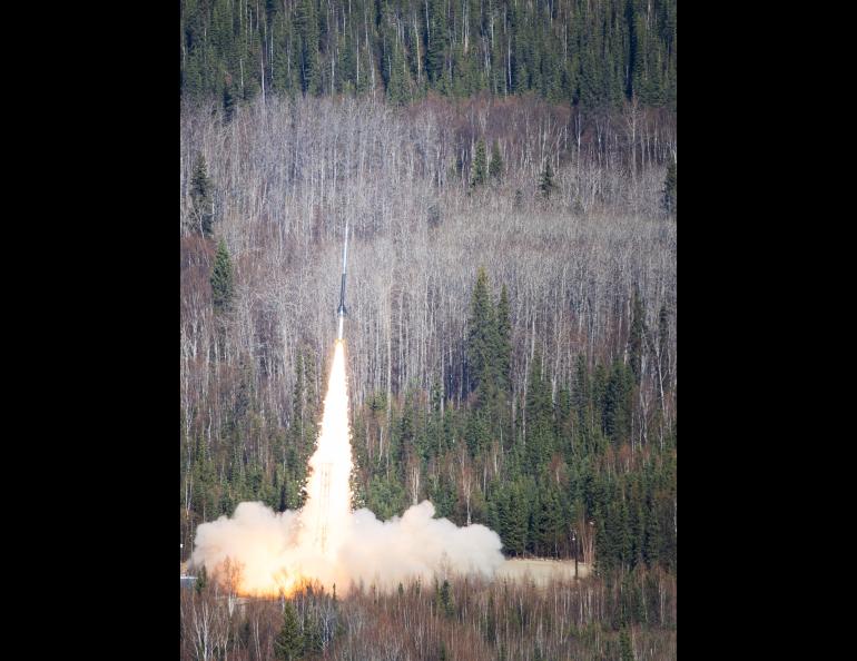 A NASA sounding rocket carrying the FOXSI-5 mission lifts off from Poker Flat Research Range north of Fairbanks at 11:23 a.m. Thursday, May 14, 2026. A bright plume of flame and smoke rises from the launch pad as the rocket climbs above a dense forest of spruce and leafless birch trees.