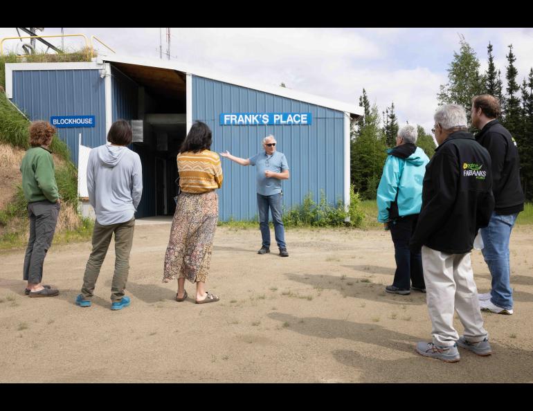 Dave Hill explains the function of the Blockhouse Complex during a public walking tour of Poker Flat Research Range on June 13, 2024. Hill gestures toward the blue blockhouse building as tour participants stand outside listening on a gravel roadway surrounded by trees and grassy berms.
