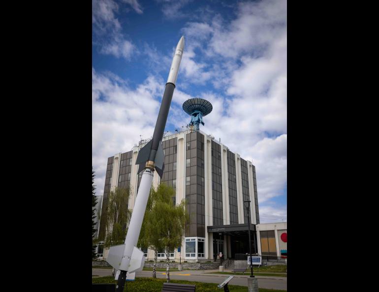Visitors on the Geophysical Institute summer tour will learn about the large blue satellite antenna atop the Elvey Building and the sounding rocket installation displayed on the lawn outside. The photo shows the rocket model rising in the foreground in front of the building under a partly cloudy sky