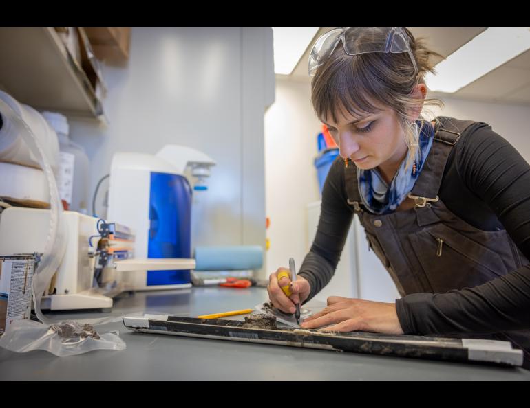 Assistant research professor Shannon Hibbard of the Desert Research Institute leans over a lab bench, carefully labeling and packaging permafrost core samples for storage. Wearing safety glasses and work overalls, she uses a marker to note details on a split core, with equipment in the background.
