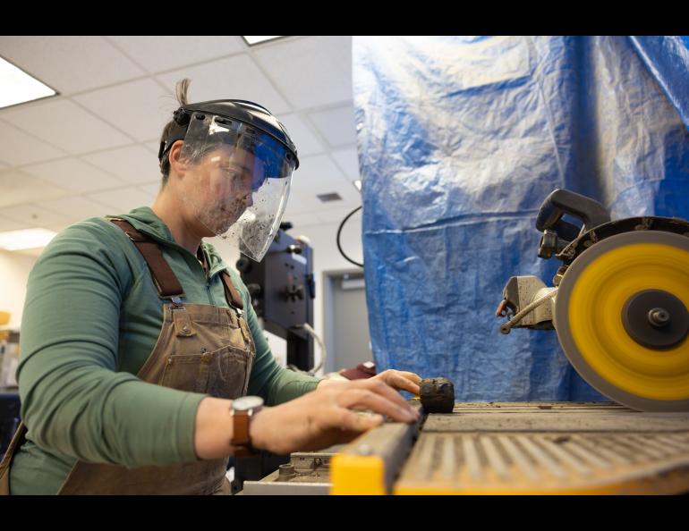UAF research assistant professor Louise Farquharson wears a face shield and apron while cutting a dark core sample on a spinning saw in a UAF laboratory. The close-up shows her guiding the sample carefully along the blade, with equipment and a blue protective curtain in the background.