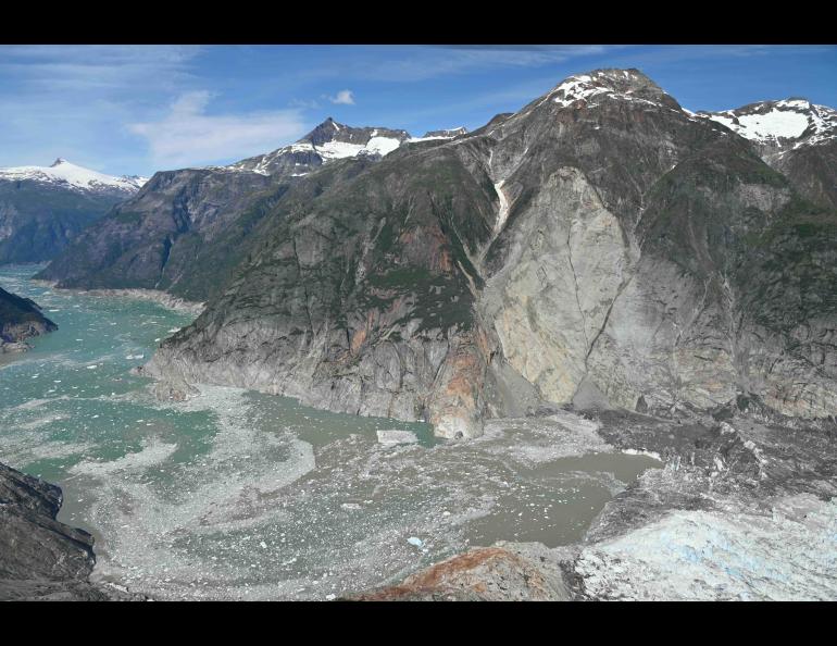 A wide aerial view shows the landslide scar, descending from a steep mountainside into the fjord near a glacier terminus. Gray rock and debris cut through the center of the mountain, reaching the water below, where floating ice and sediment spread across the fjord.