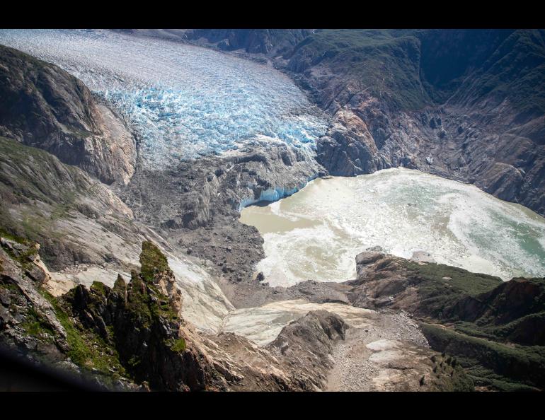 An aerial photograph looks down into the steep-walled Tracy Arm fjord in Southeast Alaska. In the foreground, a broad landslide scar cuts down a rocky mountainside toward the water, leaving exposed gray and tan rock and scattered debris.