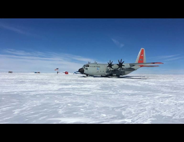 A New York Air National Guard LC-130 Skibird cargo aircraft sits on a snow runway atop the Greenland Ice Cap under a clear blue sky. The ski-equipped aircraft, operated by the 109th Airlift Wing, is parked on the windswept ice with a small field camp, flags and support structures visible.