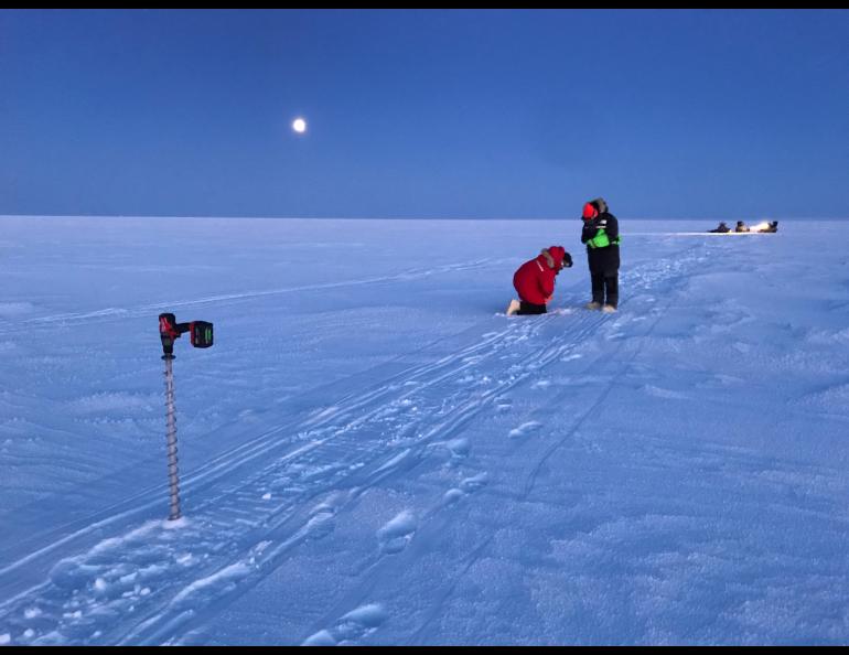  Two researchers in heavy winter clothing kneel and stand on the snow-covered surface of Teshekpuk Lake as they conduct ice measurements under a moonlit Arctic sky. An ice auger stands in the foreground near snowmachine tracks, while additional team members and equipment are in the distance. 