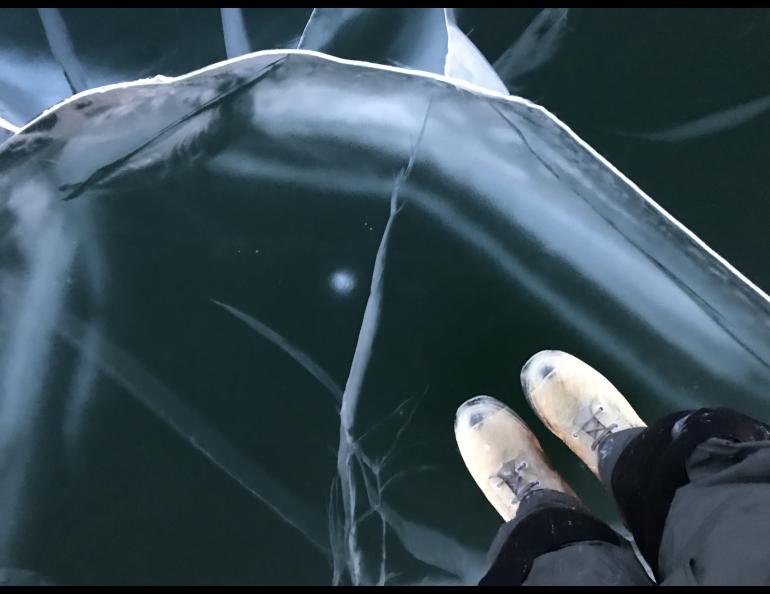 A research team member stands on the smooth, transparent surface of Teshekpuk Lake's clear ice, known as black ice. The person's boots are visible against the dark ice, which is crisscrossed by pale fractures and trapped air features. It is actually clear ice, revealing the dark of the deep.