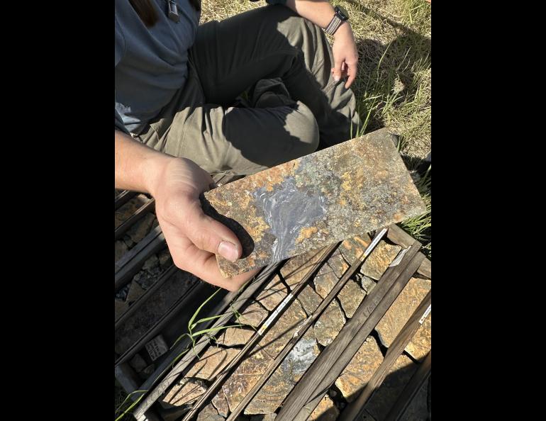 Lee Ann Munk of the University of Alaska Fairbanks Geophysical Institute holds an exploration rock core containing gallium- and silver-rich galena above boxes of core samples. Munk had recently visited Alaska Silver’s exploration site in western Alaska; the company is project partner.
