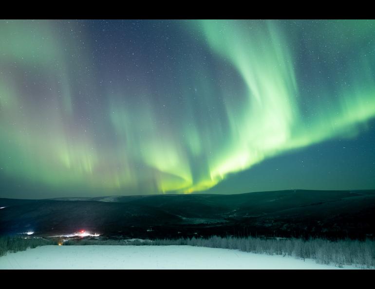 Green aurora curtains stretch across the night sky above snow-covered hills at Poker Flat Research Range north of Fairbanks in late January 2025. Facility lights glow in the valley below.