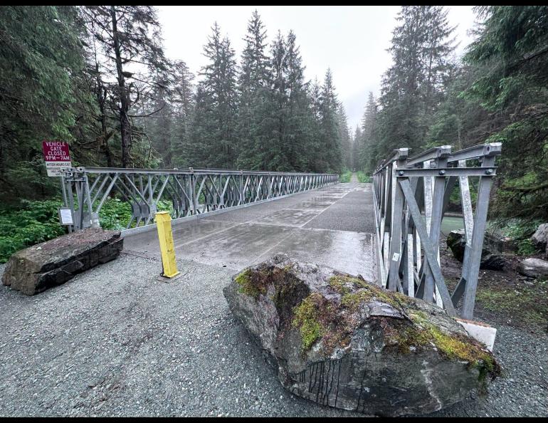Rainy silver metal pedestrian bridge crosses Montana Creek in Juneau, surrounded by dense evergreen forest. The wet metal bridge deck leads into the trees, with the creek visible below on the right.