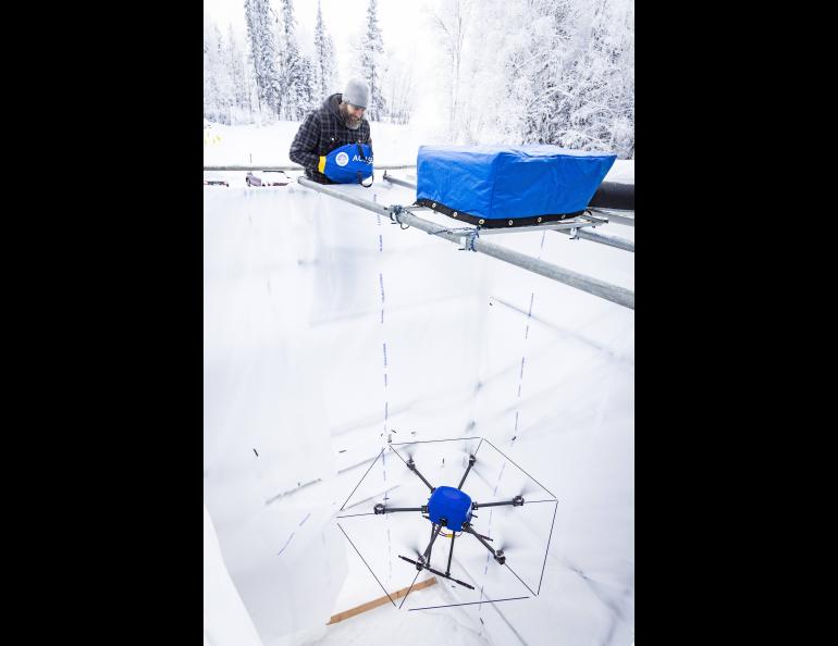 ACUASI pilot Matthew Westhoff flies a large multirotor drone inside the new University of Alaska Fairbanks icing tower in February 2025. Westhoff stands on a metal platform above the white plastic-lined test chamber, surrounded by snow-covered ground and heavily frosted forest, while the drone hover