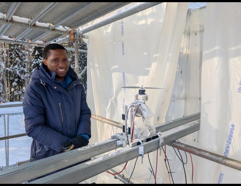 Doctoral student Morakinyo Labiran stands at ACUASI’s ice tower beside his planned test item, a small rotor assembly mounted on metal rails and surrounded by wiring. He wears a heavy blue winter coat and gloves inside the translucent plastic-lined, scaffold-supported enclosure.