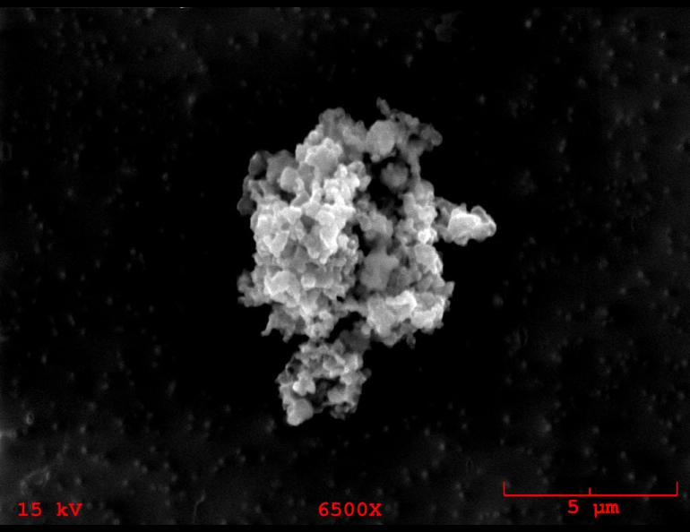 A grain of cosmic dust collected from Earth’s stratosphere appears as an irregular, lumpy cluster of gray and white material against a black background in this highly magnified image. The grain is about five micrometers across, approximately the size of a human red blood cell.