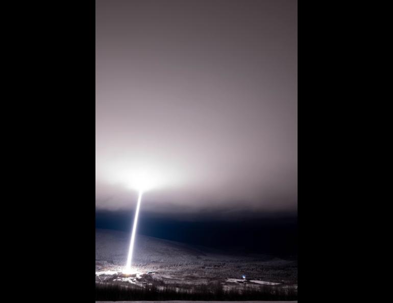 A two-stage NASA Black Brant IX sounding rocket creates a brilliant white exhaust trail that rises from the brightly illuminated launch site through the dark sky, lighting up low clouds above the snow-covered hills and forest.