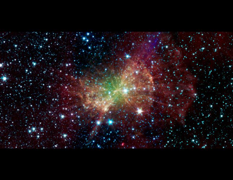 The Dumbbell Nebula glows in infrared light in this 2011 image from NASA’s Spitzer Space Telescope. A broad, irregular cloud of green, yellow and orange gas fills the center, surrounded by wispy red clouds against the blackness of space. Hundreds of bright blue-white stars are scattered throughout.