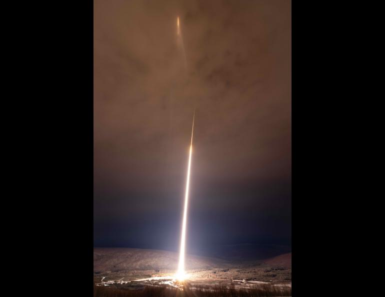 A two-stage NASA Black Brant IX sounding rocket creates a brilliant white exhaust trail that rises from the brightly illuminated launch site above snow-covered hills and forest, extending into orange-tinted clouds, where the rocket appears as a small bright streak.