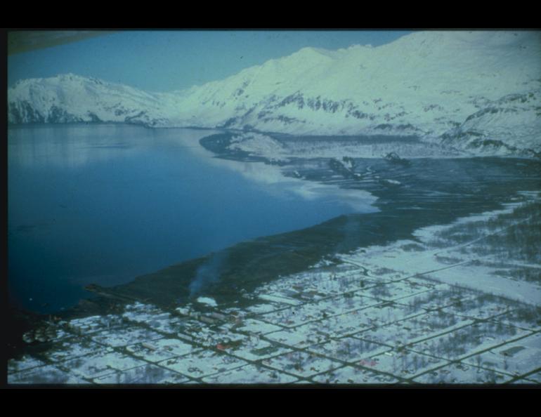 Aerial view of Valdez, Alaska, after the 1964 magnitude 9.2 earthquake, looking across the snow-covered community and shoreline toward steep, snow-covered mountains. Along the waterfront, a broad dark, snow-free area marks the extent of tsunami flooding, contrasting with the surrounding snow.
