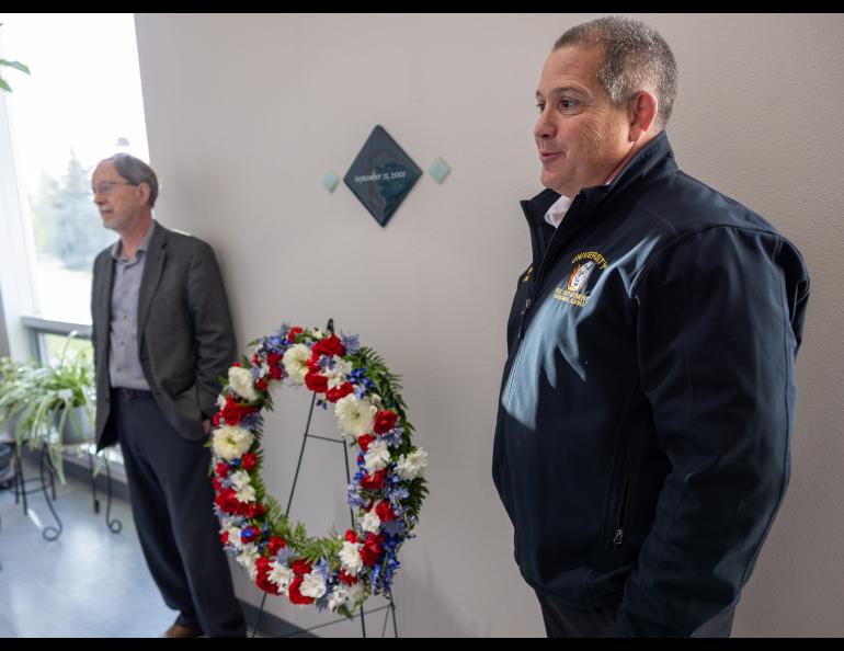 Two men stand on either side of a wreath with red, white and blue flowers on a stand in the Elvey hall.