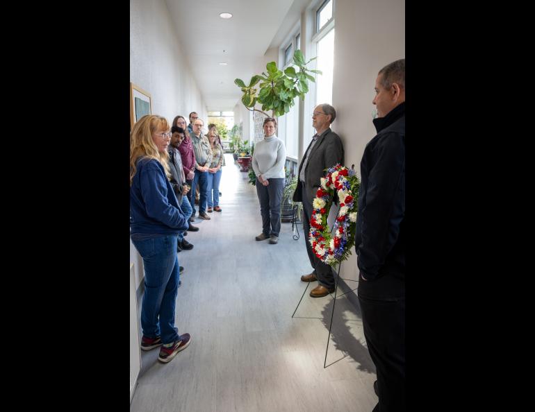 A small group of people are gathered in the Elvey Building's Bakerville Plant Hall l near a wreath of red, white and blue flowers. 