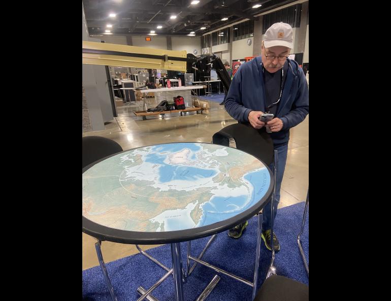 Rod Boyce checks his messages next to a table with a map of the Arctic Region GI Design Services created to put on top of the round tables in the UAF booth at AGU24. Photo by Becky LIndsey