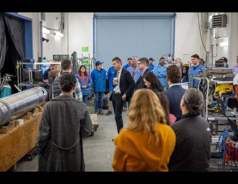 A group of people standing in a semi-circle in a large room looking at part of a rocket lying horizontally in a stand