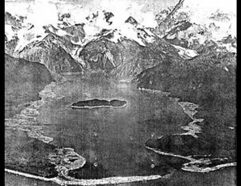 The central portion of T-shaped Lituya Bay with Cenotaph Island in its center. Mt. Crillion, 12, 726 feet high, towers over other peaks surrounding the bay. Treeless areas of the shore were wiped clean by the wave which swept down the bay on July 10, 1958. Photo by Byron Hale.