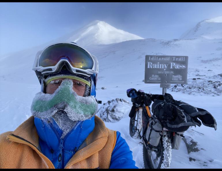 Peter Delamere poses in front of a sign in Rainy Pass in the Alaska Range during the 2024 Iditarod Trail Invitational race. On his face is a “nose-hat” invented by Fairbanks athlete Shalane Frost. Photo by Peter Delamere.