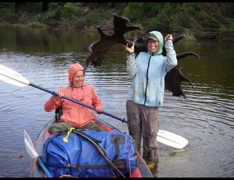 Molly Yazwinski holds a 3,000-year-old moose skull with antlers still attached found in a river on Alaska’s North Slope. Her aunt, Pam Groves, steadies an inflatable canoe. Photo by Dan Mann.