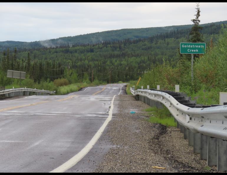 A bumpy asphalt road goes through a forest. A green road sign next to it says "Goldstream Creek"