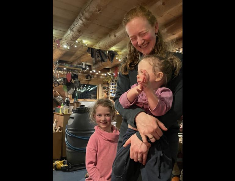 Jenna Jonas holds her daughter Juniper while her other daughter Celia looks on. Jenna and David Jonas hosted Ned and Forest at their Tanana River homestead on the first night of the bikers’ trip. Photo by Ned Rozell.