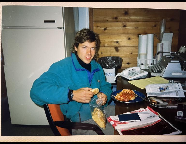 A young man in a green fleece sweater sits behind a desk while eating a plate of spaghetti and holding a piece of bread. An old typewriter sits on the desk in the background.
