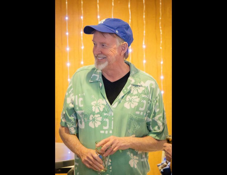A person with a blue cap and green shirt holding a pint glass stands in front of a yellow background