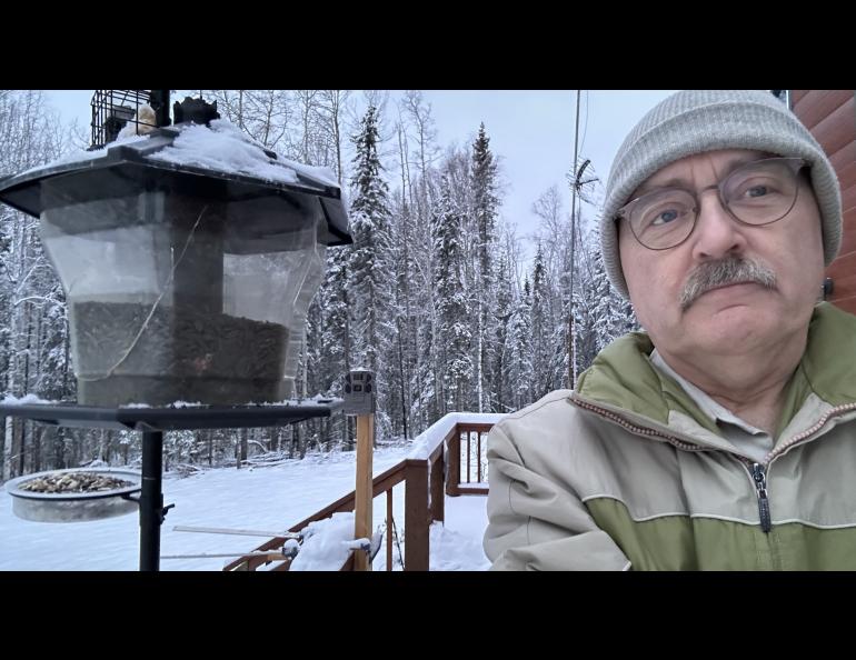 A person stands in front of a bird feeder against a snowy forest background.