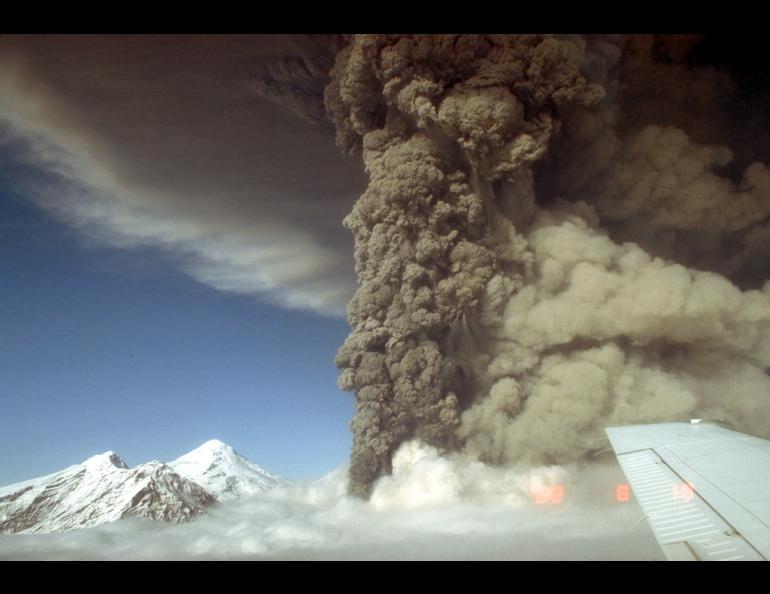 A towering dark gray ash plume rises from Mount Spurr’s Crater Peak vent during the Aug. 18, 1992, eruption. A lighter tan cloud from pyroclastic flows spreads to the right, while the snow-covered 11,070-foot summit lava dome complex is visible at left beneath a blue sky. Part of an aircraft wing ap