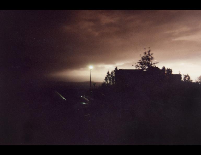 A dark brown-gray ash cloud from Mount Spurr spreads over Anchorage on Aug. 18, 1992, dimming the late-afternoon sky above Rod Boyce’s condominium near Cook Inlet. A bright band remains near the horizon while buildings, trees and parked cars appear in silhouette and a streetlight glows beneath the a